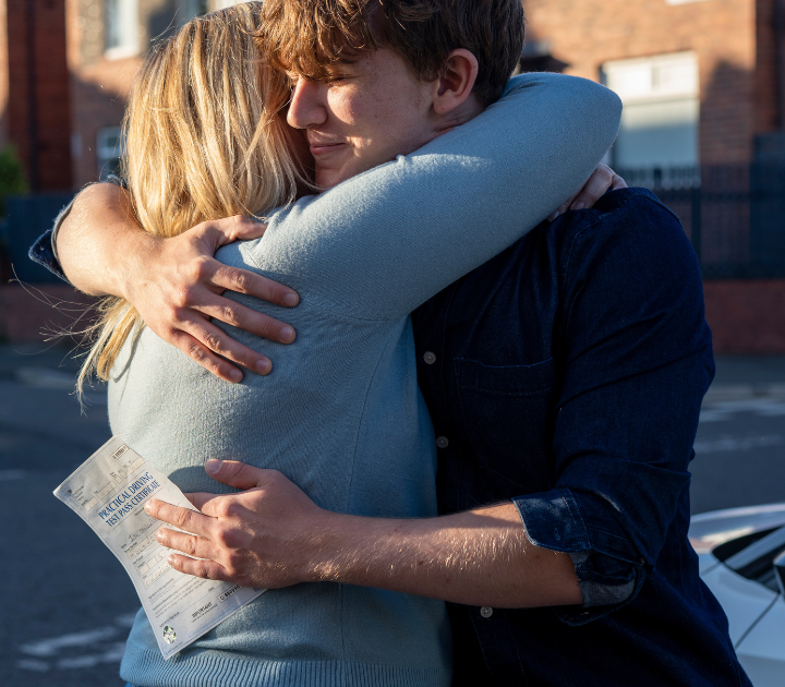 student hugging a woman whilst holding their practical driving test pass certificate after passing their driving test